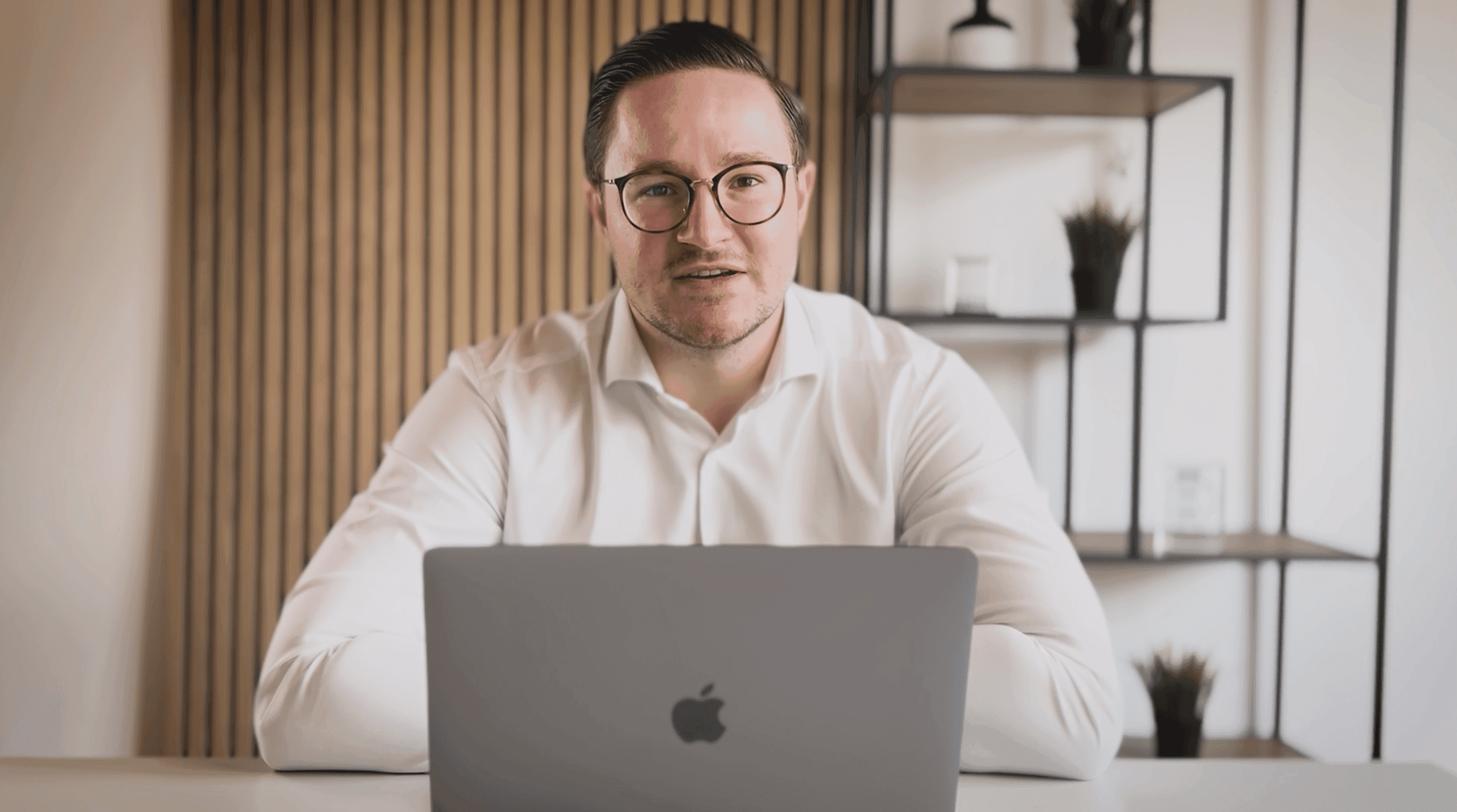 Erik Schmezer mit Brille und weißem Hemd am Schreibtisch vor einem aufgeklappten MacBook, im Hintergrund eine Holzlamellenwand