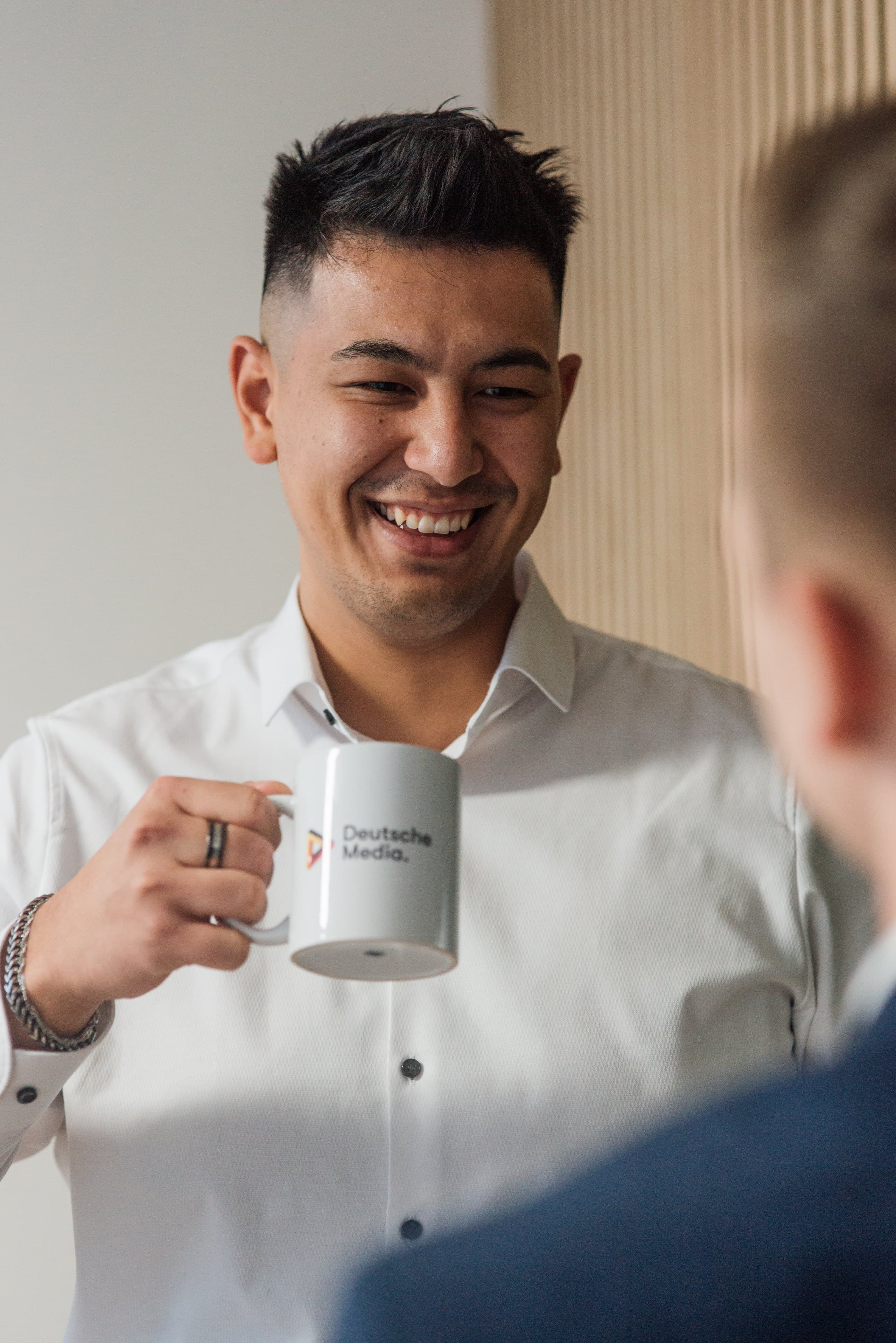 Marco Kaiser im weißen Hemd, lachend im Gespräch mit einem Kollegen, in der Hand eine Tasse mit dem Logo von Deutsche Media